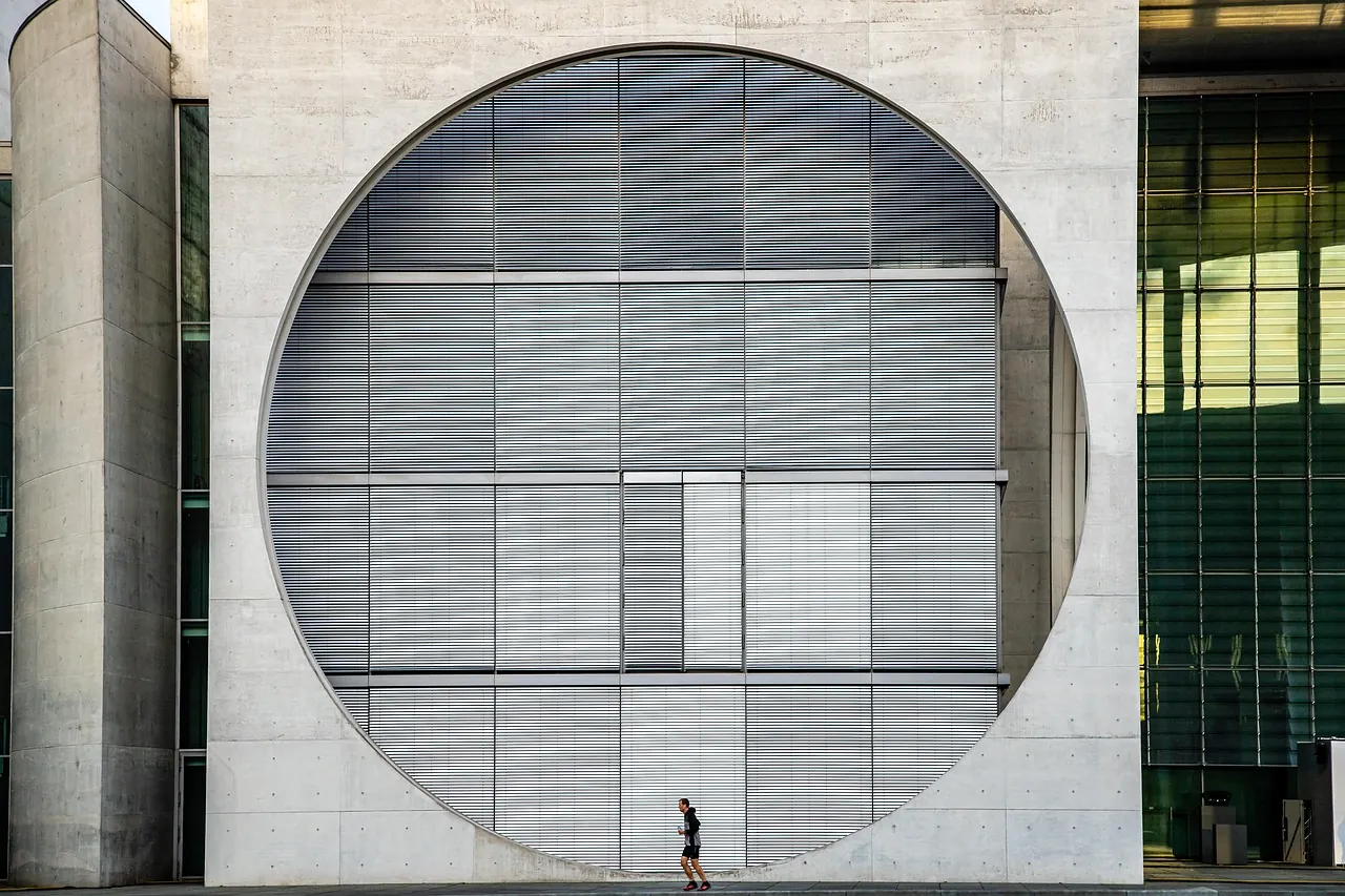 A monumental circular window in a concrete civic facade, a person walking past